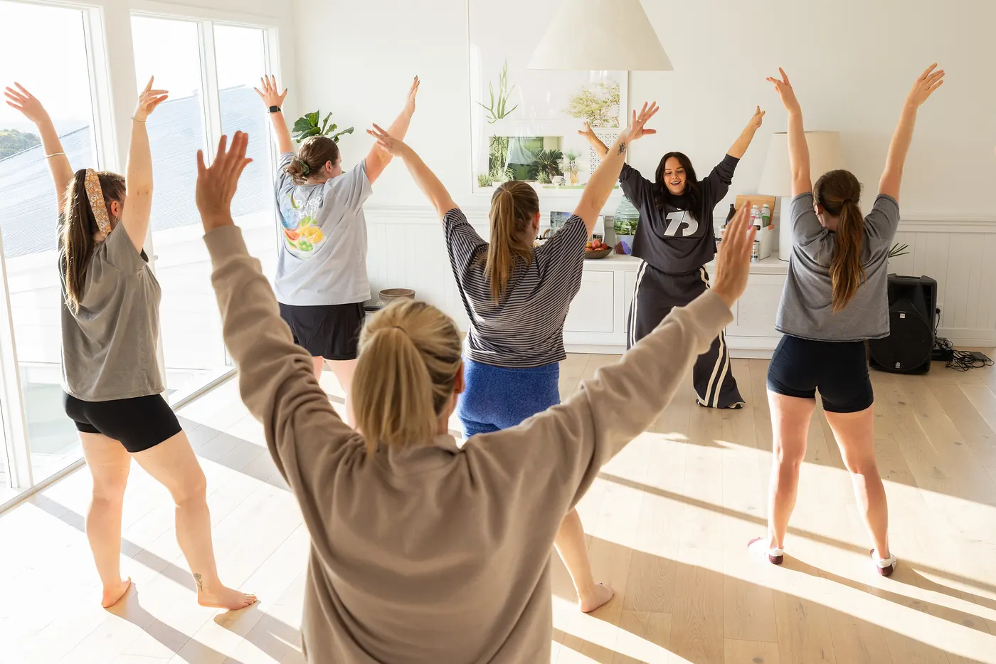 Women in a sunlit studio throwing their arms up during a dance class