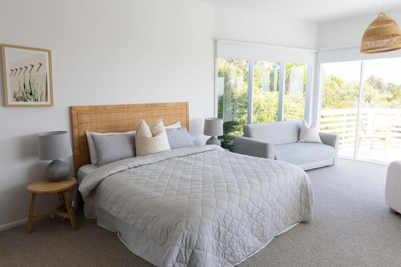 A bright bedroom with a rattan bedhead, a grey quilt and doors to a balcony